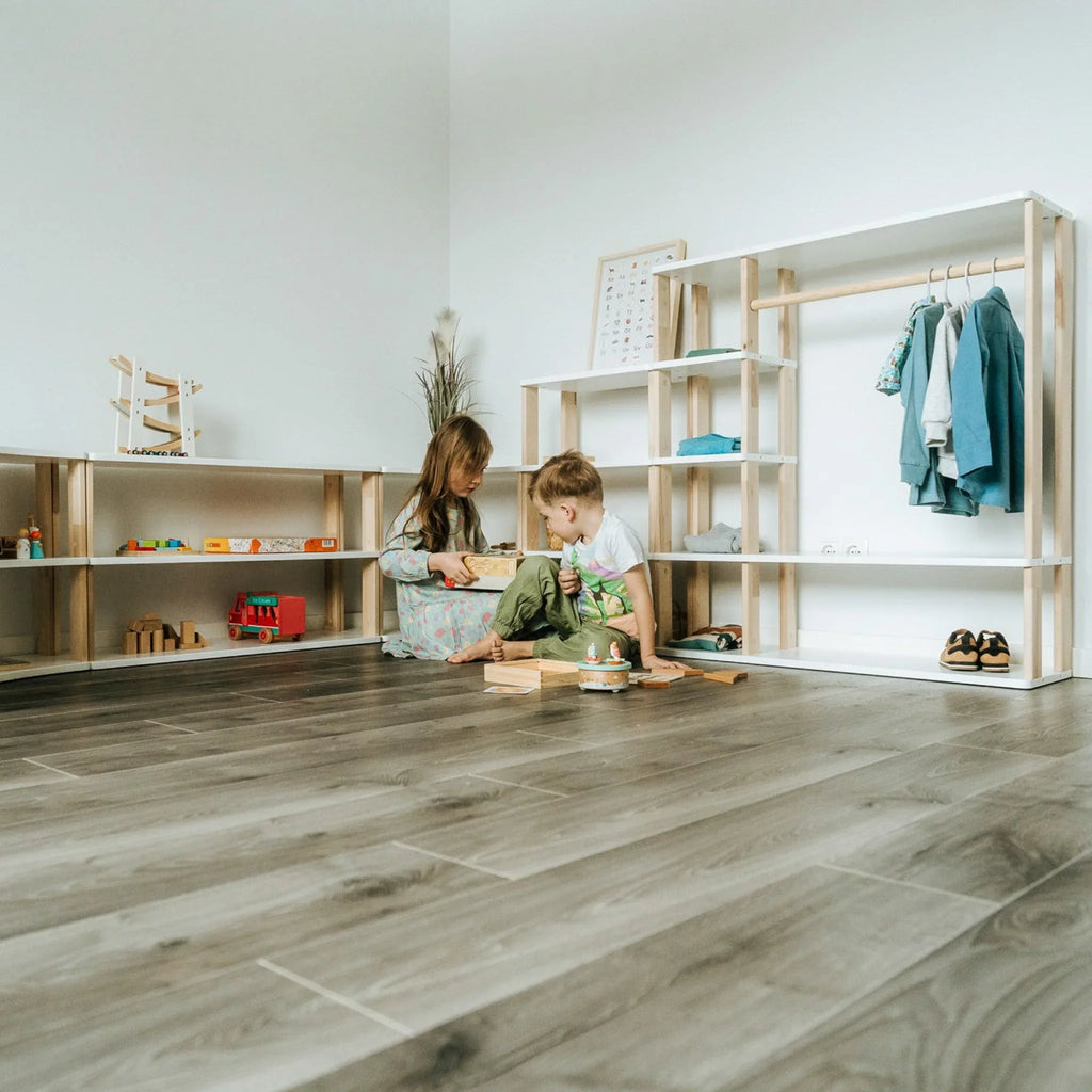 Two children playing near a wooden Montessori bookshelf with open shelves and hanging space