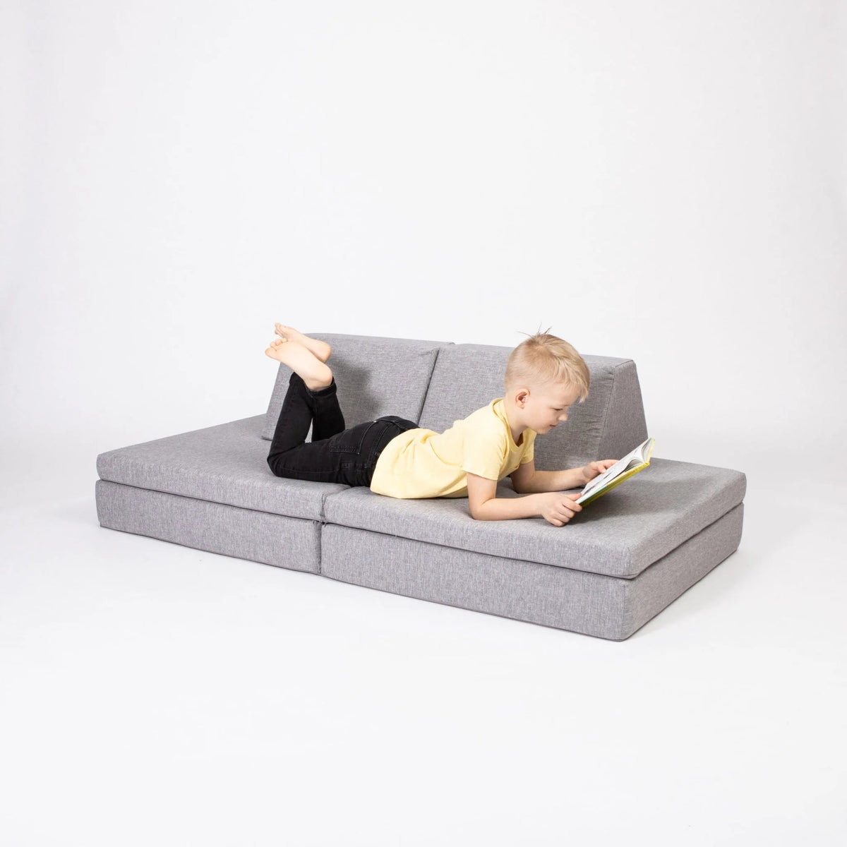Child reading a book on a gray sectional sofa against a white background