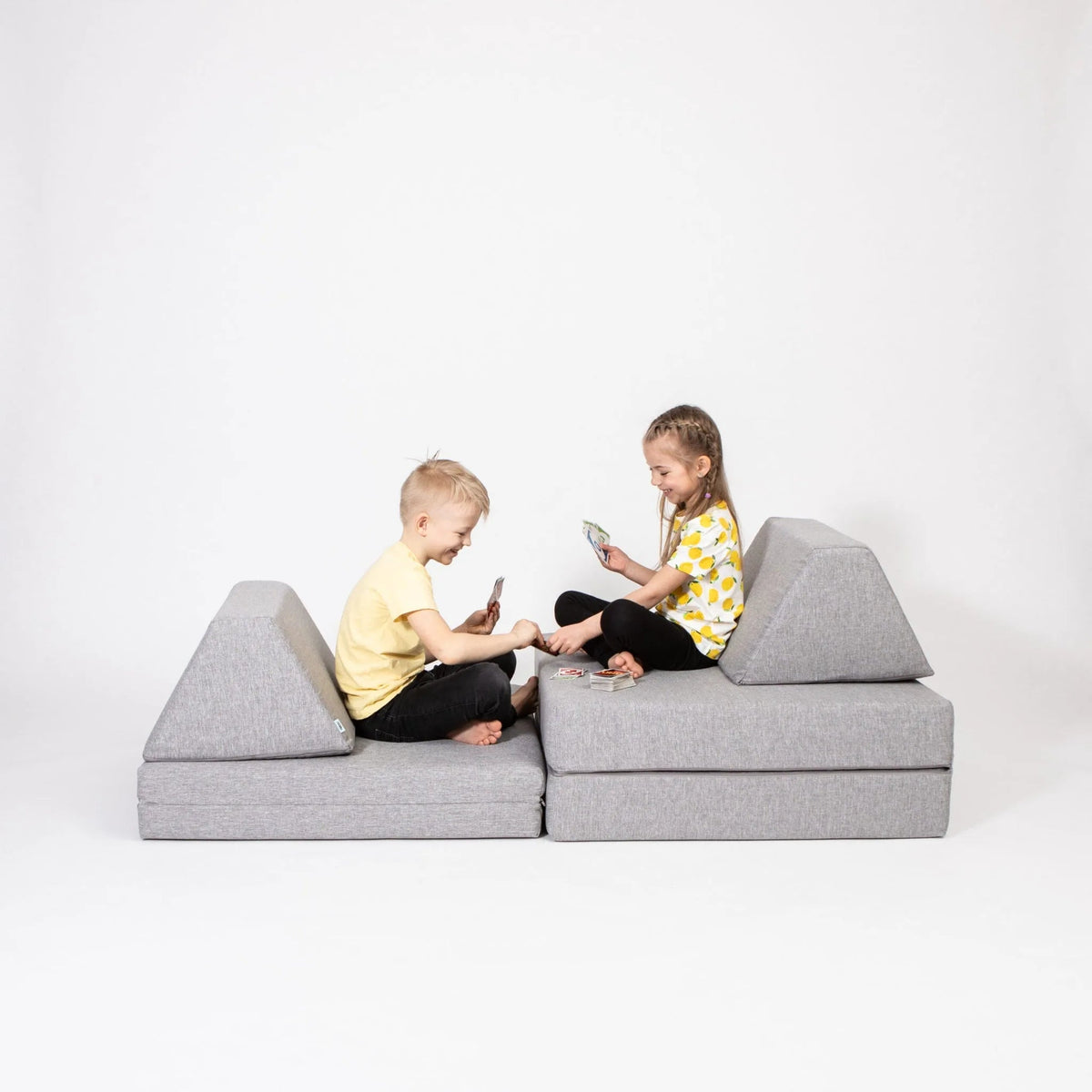Two children sitting on a gray wedge-shaped sofa against a white background