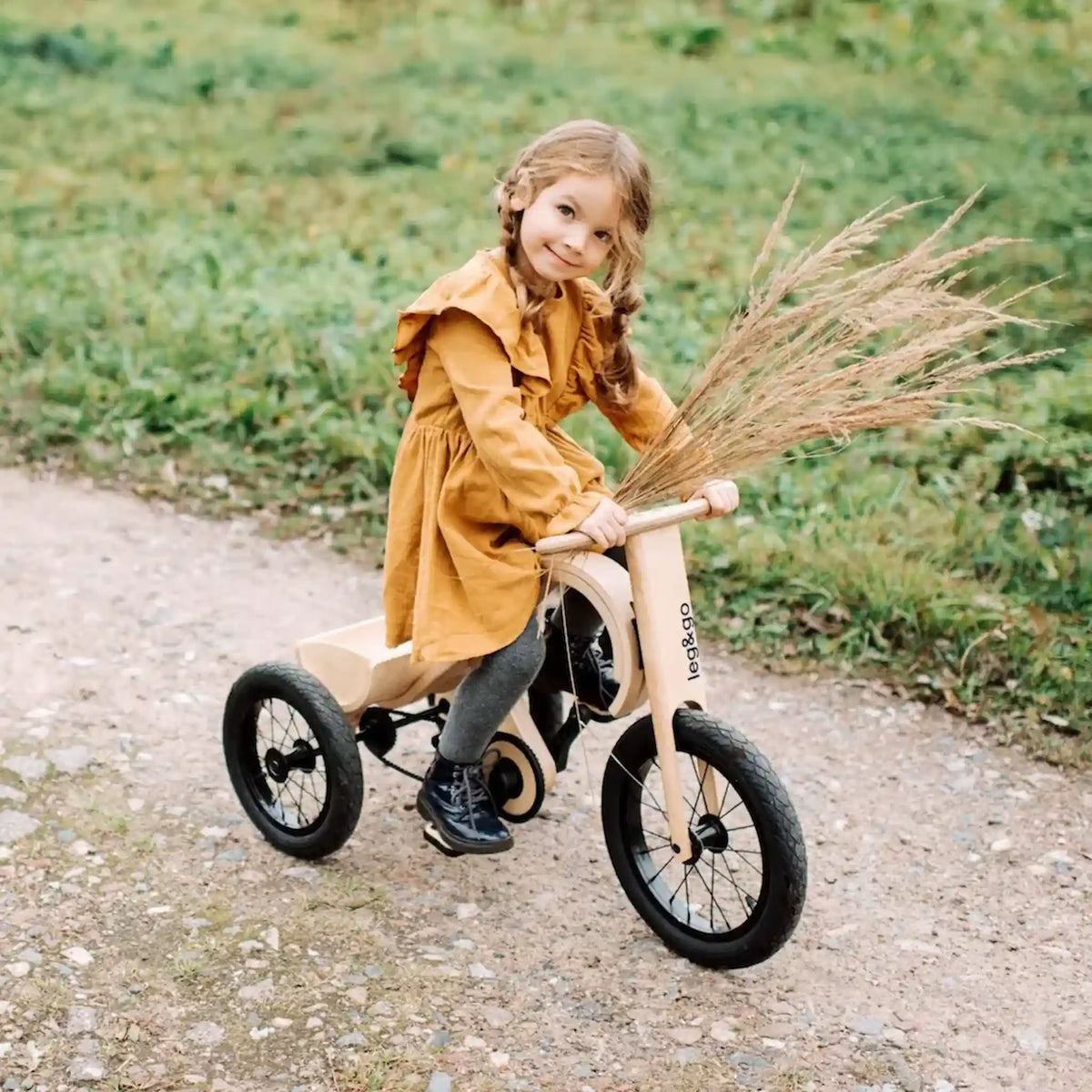 Child in a yellow coat riding a balance bike with a broom in a natural setting