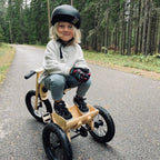 Child riding a wooden bike on a paved road with a forest in the background